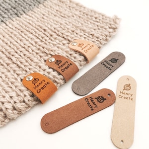 May include: Five leather stitch markers with the text "Henry Create" and a small yarn ball design. The stitch markers are in various shades of brown, beige, and grey.
