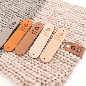 May include: Six leather labels with the text "Henry Create" and a small yarn ball icon in various shades of brown, beige, and orange. The labels are arranged in a row on a beige crocheted surface.