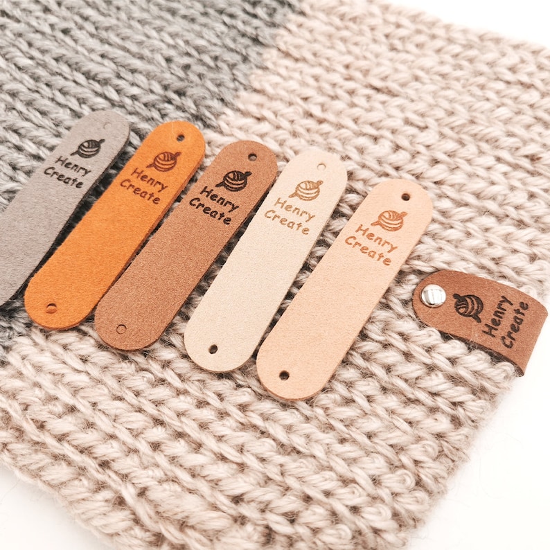 May include: Six leather labels with the text "Henry Create" and a small yarn ball icon in various shades of brown, beige, and orange. The labels are arranged in a row on a beige crocheted surface.