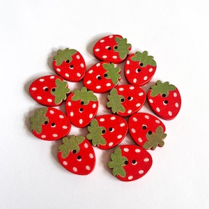 May include: A cluster of red wooden strawberry-shaped buttons with white dots and green leaf details. Each button has two holes for sewing. The buttons are scattered on a white surface.
