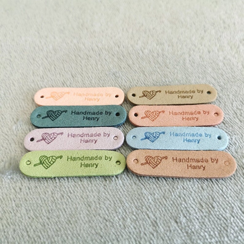 Pode incluir: Dez etiquetas de couro ovais com um design de cora&ccedil;&atilde;o e fio. Cada etiqueta diz "Handmade by Henry". As etiquetas s&atilde;o de v&aacute;rias cores, incluindo rosa, verde, azul e marrom.