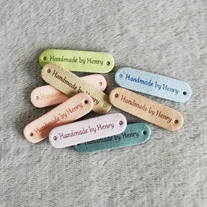 Peut inclure: Gros plan sur dix &eacute;tiquettes en cuir color&eacute;es avec le texte "Handmade by Henry" sur chacune. Les &eacute;tiquettes sont dispos&eacute;es en pile sur une surface douce et duveteuse.