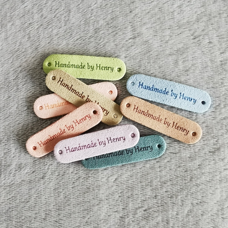 Peut inclure: Gros plan sur dix &eacute;tiquettes en cuir color&eacute;es avec le texte "Handmade by Henry" sur chacune. Les &eacute;tiquettes sont dispos&eacute;es en pile sur une surface douce et duveteuse.