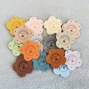 May include: A collection of 14 flower-shaped leather patches in various colours, each with a brown circle in the centre that reads "handmade with" and a small heart symbol.