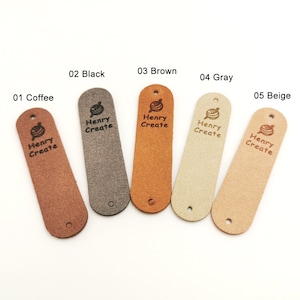May include: Five leather labels in different colours: coffee, black, brown, grey, and beige. Each label has a hole at the top and a small circle at the bottom. The labels are all rectangular and have the text "Henry Create" and a small yarn ball icon printed on them.