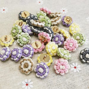 May include: A collection of round buttons featuring a gold-toned metal centre surrounded by a crocheted flower design in various pastel colours including pink, yellow, green, purple, and brown, with white thread accents throughout the petals.