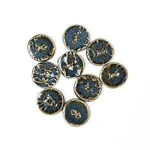 May include: A collection of ten ceramic buttons with a glossy, textured finish. The buttons are primarily blue with brown accents and feature various embossed designs. Each button has two or four holes for sewing.