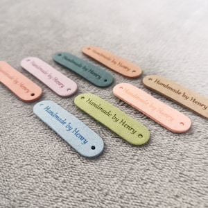 Peut inclure: Assortiment de huit &eacute;tiquettes en cuir color&eacute;es avec le texte "Handmade by Henry" en lettres blanches. Les &eacute;tiquettes sont de diff&eacute;rentes couleurs, notamment rose, bleu, vert et marron.