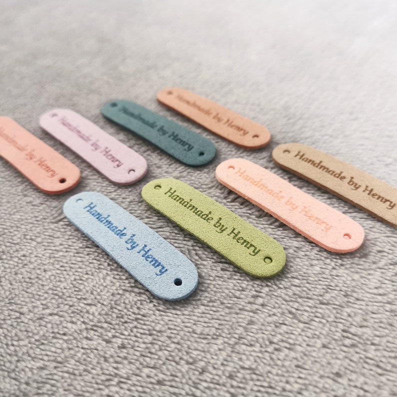 Peut inclure: Assortiment de huit &eacute;tiquettes en cuir color&eacute;es avec le texte "Handmade by Henry" en lettres blanches. Les &eacute;tiquettes sont de diff&eacute;rentes couleurs, notamment rose, bleu, vert et marron.