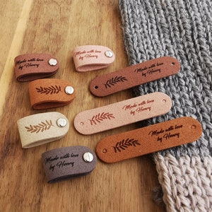 May include: Assortment of six leather labels with a leafy design and the text "Made with love by Henry" in brown lettering. The labels are in various shades of brown, beige, and pink.