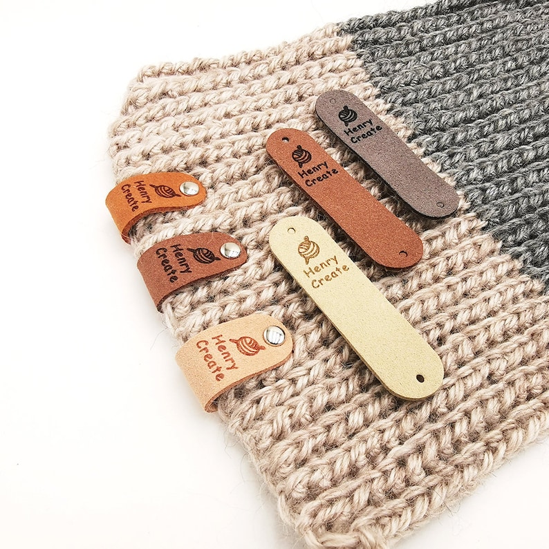 May include: Five leather tags with the text "Henry Create" and a small yarn ball design. The tags are in various colours and are attached to a crocheted fabric.