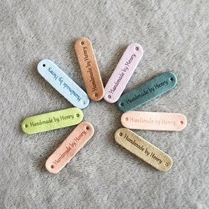 May include: Seven colourful fabric labels with rounded edges and holes. Each label has the text "Handmade by Henry" written in black.