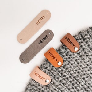 May include: Four brown leather tags with the name "HENRY" engraved on them. The tags are attached to a grey knitted hat.