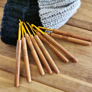 Puede incluir: Una colecci&oacute;n de ganchillos de madera con puntas de metal dorado. Cada ganchillo est&aacute; grabado con un nombre y el tama&ntilde;o, 5,0 mm o 5,5 mm. Los ganchillos est&aacute;n colocados sobre una superficie de madera, con un gorro de punto en el fondo.