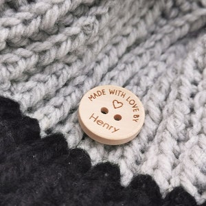 May include: A wooden button with the text "Made with love by Henry" and a small heart engraved on it. The button is on a gray and black crocheted fabric.