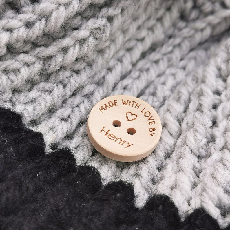 May include: A wooden button with the text "Made with love by Henry" and a small heart engraved on it. The button is on a gray and black crocheted fabric.
