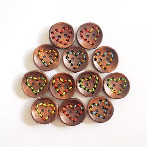 May include: A selection of wooden buttons featuring a heart design. The brown buttons have embroidered hearts in red, orange, yellow, green, and blue thread. Each button is round with two holes for sewing, ideal for craft projects.