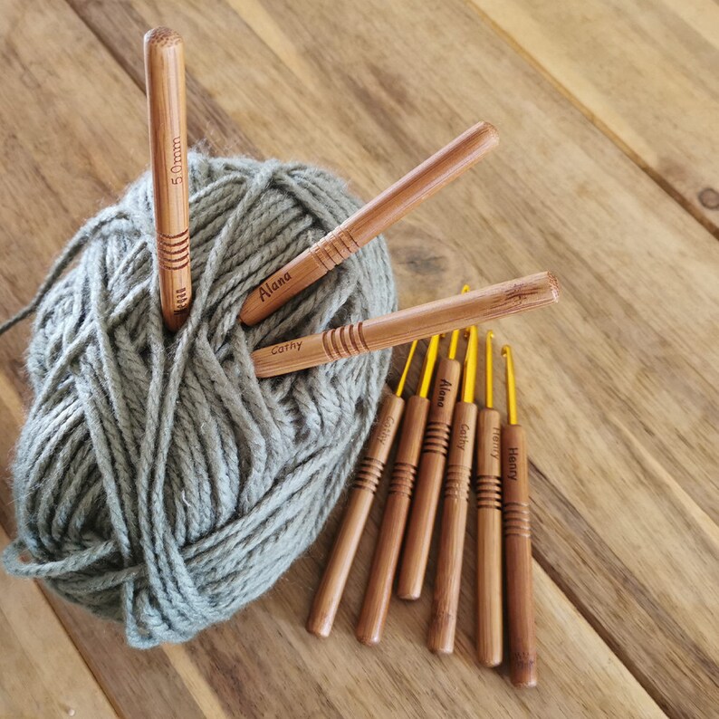 Puede incluir: Un conjunto de ganchillos de bamb&uacute; con puntas doradas, algunos personalizados con nombres, y una madeja de lana gris. Un ganchillo est&aacute; marcado con "5.0mm". Los ganchillos est&aacute;n colocados sobre una superficie de madera.
