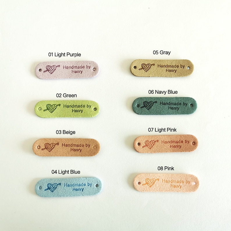 Pode incluir: Oito etiquetas de couro de diferentes cores com um design de cora&ccedil;&atilde;o e flecha e o texto "Handmade by Henry". As etiquetas s&atilde;o de cor roxo claro, verde, bege, azul claro, cinza, azul marinho, rosa claro e rosa.