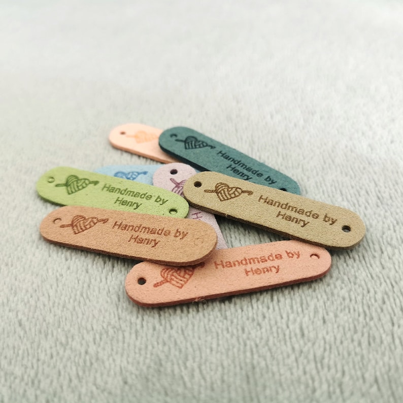 Pode incluir: Um close-up de v&aacute;rias etiquetas de couro ovais com um design de cora&ccedil;&atilde;o e flecha. As etiquetas s&atilde;o de v&aacute;rias cores, incluindo verde, azul, rosa e marrom. Cada etiqueta tem o texto "Handmade by Henry" gravado nela.