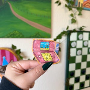 May include: A pink sticker in the shape of a retro handheld video game console. The screen displays a colorful landscape with flowers. The buttons and directional pad are yellow. The sticker is held in a hand with a dark green nail.