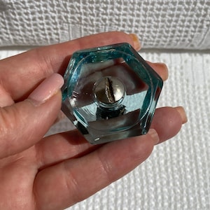 Pode incluir: Um puxador de armário hexagonal de vidro azul claro com um parafuso prateado no centro. O puxador é mantido contra um fundo branco texturizado. Este hardware decorativo é adequado para restauração de móveis ou projetos DIY.
