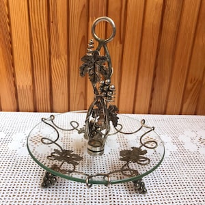 May include: Silver-tone glass serving dish with a grapevine design. The dish has a round glass base with a silver-tone grapevine design around the edge. The dish has a silver-tone handle with a grapevine design.