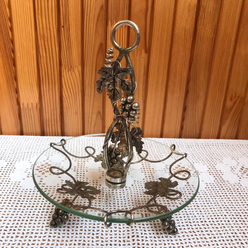 May include: Silver-tone glass serving dish with a grapevine design. The dish has a round glass base with a silver-tone grapevine design around the edge. The dish has a silver-tone handle with a grapevine design.
