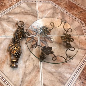 May include: A glass serving tray with a metal base featuring a grapevine design. The base is decorated with gold-colored leaves and grapes.
