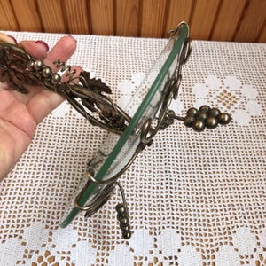 May include: A silver metal grapevine design serving tray with a glass top. The tray is shaped like a circle and has a decorative grapevine design around the edge. The tray is perfect for serving snacks, appetizers, or desserts.