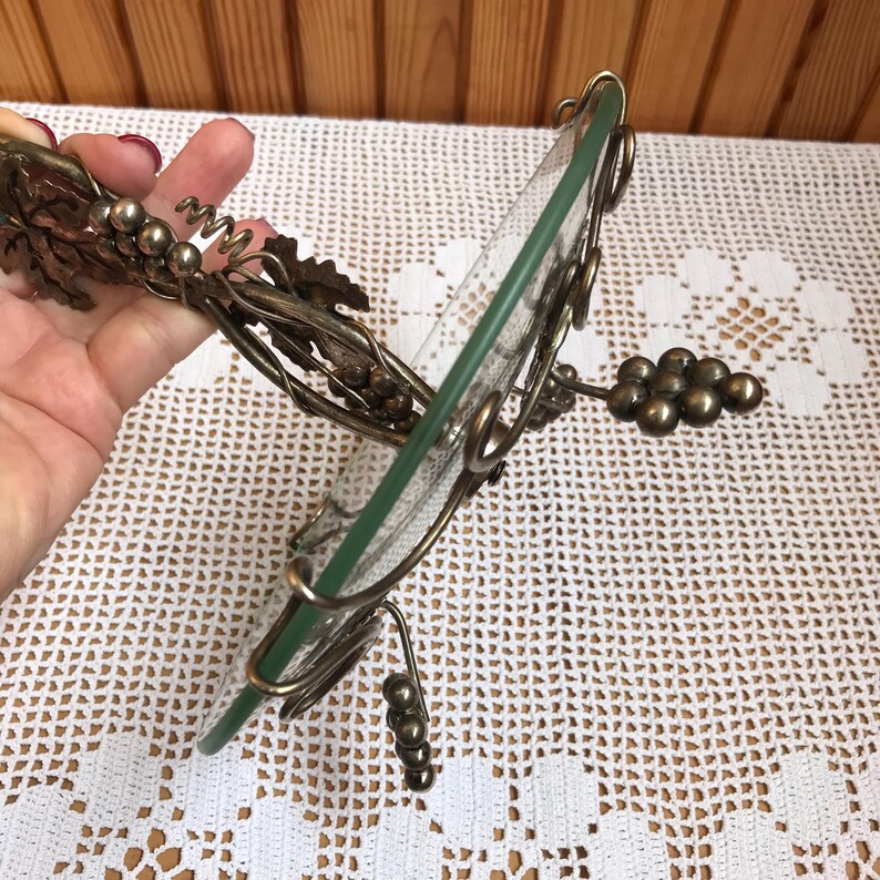 May include: A silver metal grapevine design serving tray with a glass top. The tray is shaped like a circle and has a decorative grapevine design around the edge. The tray is perfect for serving snacks, appetizers, or desserts.