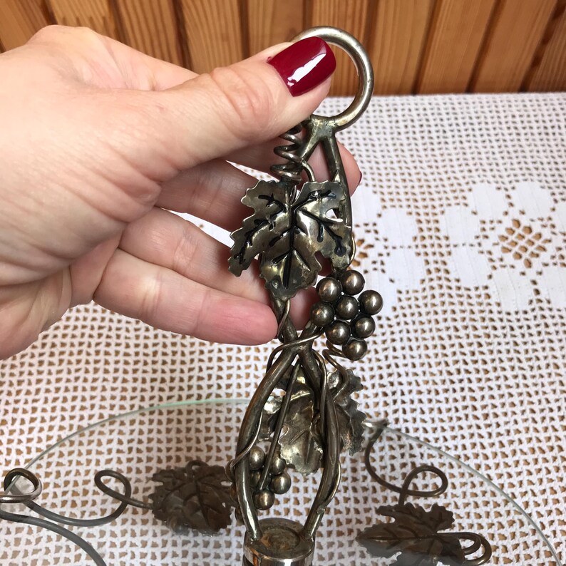 May include: Silver-toned metal serving dish with a glass base and a grapevine design. The dish has a handle with a loop at the top.