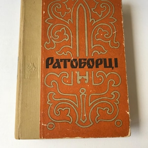 Puede incluir: Un libro antiguo de tapa dura con una cubierta naranja que presenta un intrincado diseño dorado y el título "Ратоборцы" en letras negras.