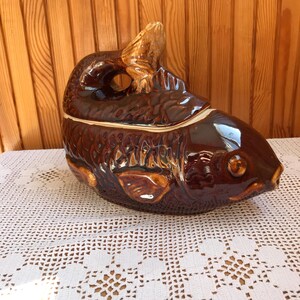 Könnte beinhalten: Eine braune, fischförmige Servierschale aus Keramik mit Deckel. Die Schale hat eine glänzende Oberfläche und detaillierte Schuppen. Der Schwanz und die Flossen des Fisches sind erhöht. Die Schale steht auf einer weißen Spitzentischdecke.