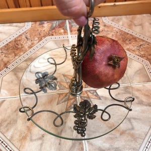 May include: A silver metal fruit stand with a glass plate and a pomegranate on top. The stand is decorated with grape vines and leaves.