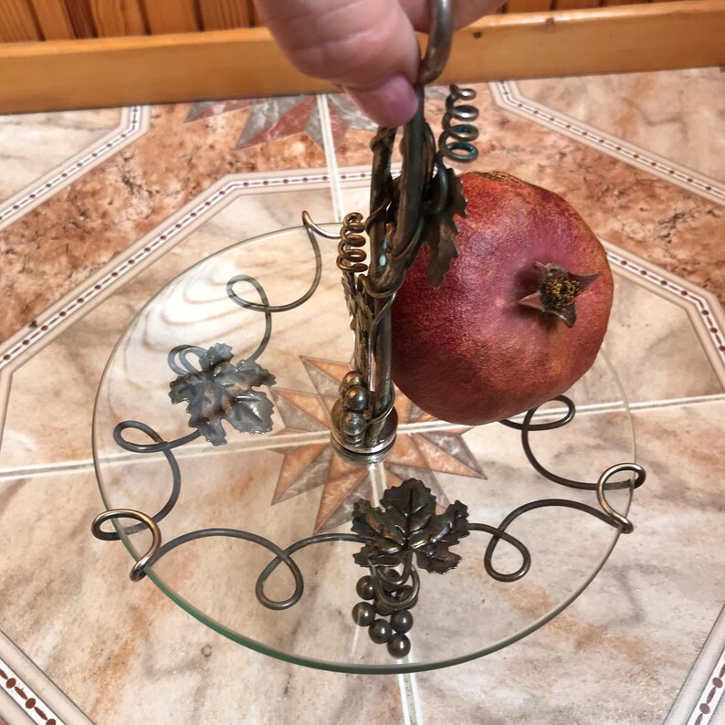 May include: A silver metal fruit stand with a glass plate and a pomegranate on top. The stand is decorated with grape vines and leaves.