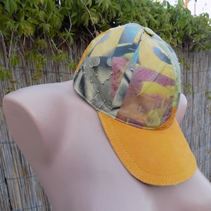 Op de afbeelding: Een gele baseballcap met een veelkleurig patchwork-ontwerp. De cap heeft een gele klep en een kroon met verschillende kleuren en patronen, waaronder groen, blauw en roze. De kroon is versierd met zilveren studs.