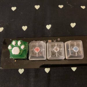 Könnte beinhalten: Eine schwarze Tastatur mit drei transparenten Tastenkappen und einer grünen Tastenkappe mit einem weißen Pfotenabdruck. Die Tastenkappen haben ein Kreuz, ein Pluszeichen und einen Kreis mit einem Punkt in der Mitte.