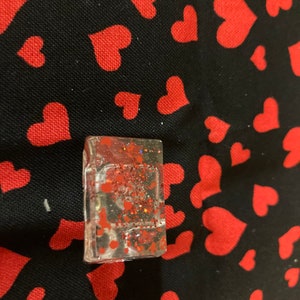 May include: A small, clear, rectangular resin charm with red glitter and red hearts inside. The charm is sitting on a black fabric background with red hearts.