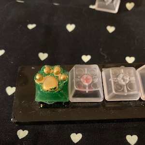 Könnte beinhalten: Eine grüne und goldene Tastaturkappe mit Pfotenabdruck und Glitzer. Die Tastaturkappe befindet sich auf einer schwarzen Tastatur mit anderen Tastaturkappen.