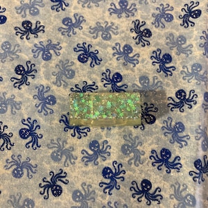 May include: A close-up of a light brown rectangular box with a glittery green surface. The box is covered in small green stars and is sitting on a white fabric background with a blue octopus pattern.