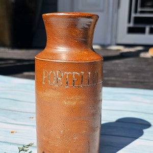 Puede incluir: Un tarro de cerámica marrón con la palabra "PORTELIO" escrita en la parte delantera. El tarro está sobre una superficie de madera azul claro.