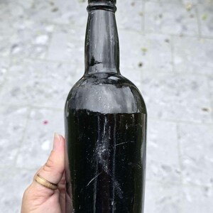 May include: A dark, antique glass bottle with a rounded body and a long neck. The bottle is black and appears to be empty. The bottle is held by a person. The bottle is approximately 25 cm tall.