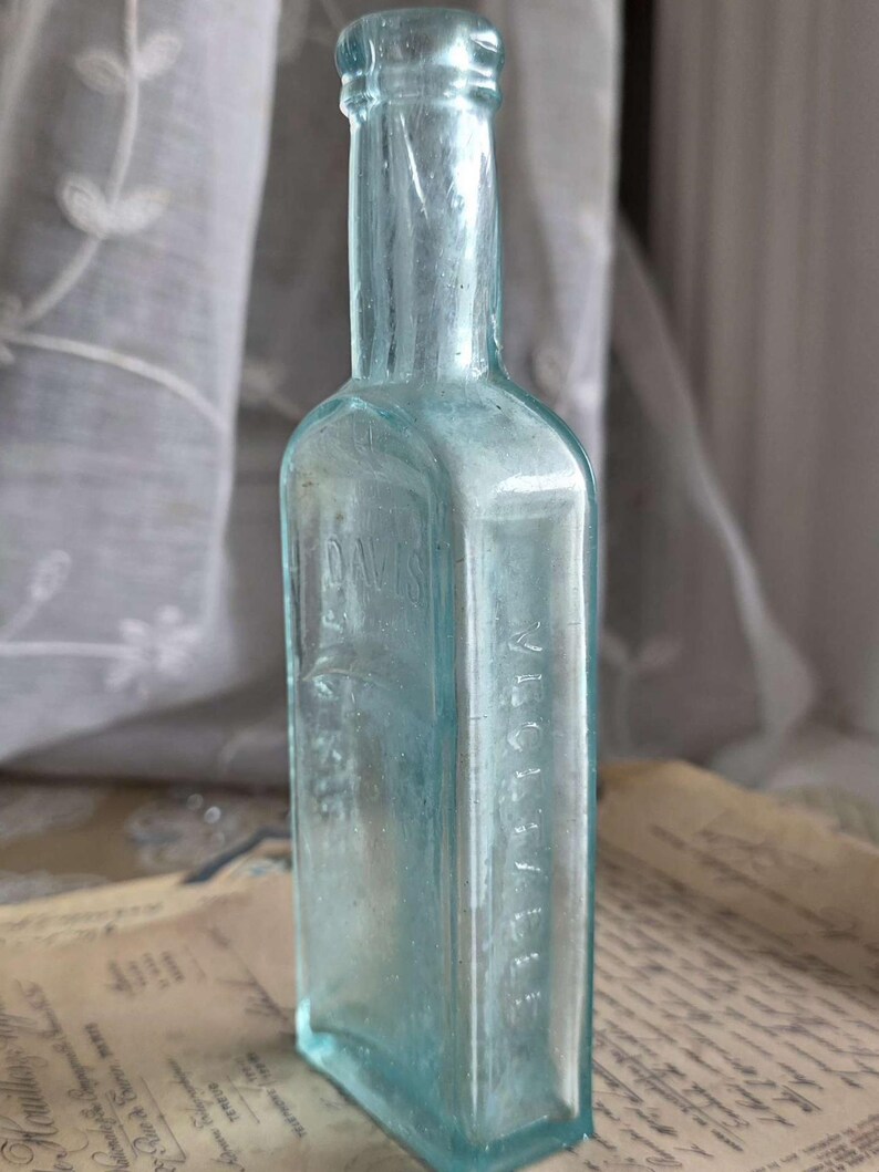 May include: A vintage, rectangular, light blue glass bottle. The bottle has a narrow neck and a slightly rounded shoulder. The text "DAVIS" is embossed on the front, and "VICHELLE" is embossed on the side. The bottle is displayed on a vintage paper.