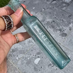 May include: A vintage, rectangular, light blue glass bottle with the embossed word "VEGETABLE". The bottle has a narrow neck and a flat base. The bottle is held by a person wearing rings.