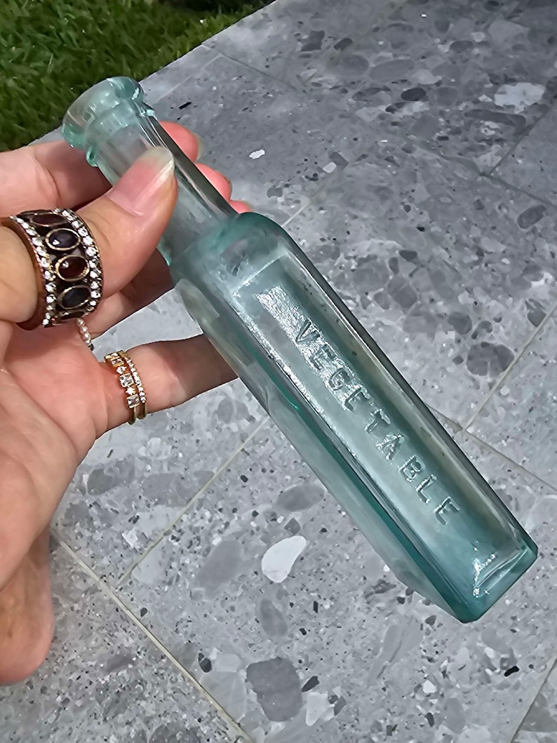 May include: A vintage, rectangular, light blue glass bottle with the embossed word "VEGETABLE". The bottle has a narrow neck and a flat base. The bottle is held by a person wearing rings.