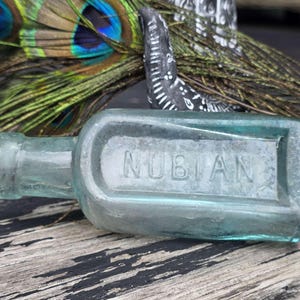 Puede incluir: Una botella de vidrio turquesa rectangular vintage con la palabra "NUBIAN" en relieve. La botella está sobre una superficie de madera desgastada, con plumas de pavo real y un elemento decorativo en el fondo.