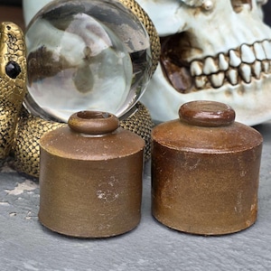 Peut inclure: Deux petits pots cylindriques en céramique marron avec des couvercles arrondis. Une statue de serpent doré berce un globe de verre transparent en arrière-plan, avec un crâne visible derrière le globe. Les pots sont sur une surface grise.