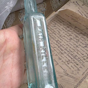 May include: A vintage, rectangular, light blue glass bottle with the embossed text "PAIN KILLER". The bottle has a narrow neck and is held against a backdrop of aged paper with handwritten script. The bottle's color is a pale, transparent blue.