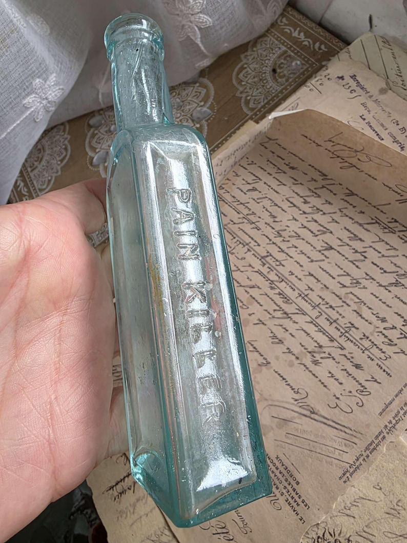 May include: A vintage, rectangular, light blue glass bottle with the embossed text "PAIN KILLER". The bottle has a narrow neck and is held against a backdrop of aged paper with handwritten script. The bottle's color is a pale, transparent blue.